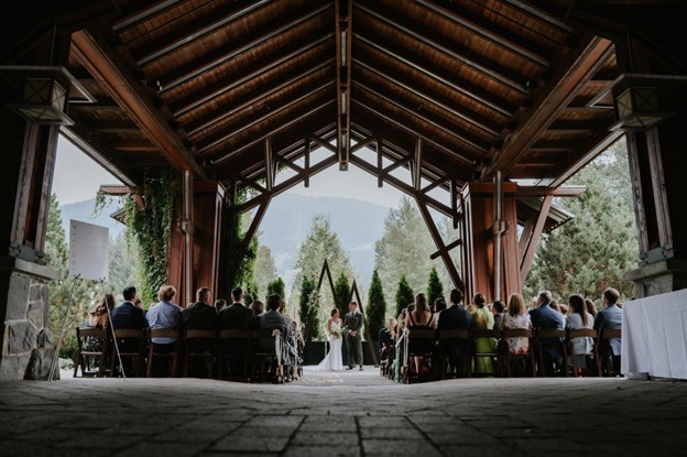 Planning a wedding in Whistler? An outdoor ceremony takes place under a wooden pavilion, with guests seated on both sides facing the couple and officiant, all surrounded by lush greenery and trees.