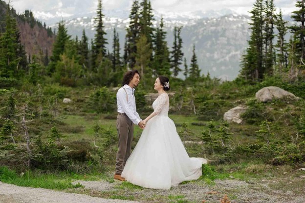 A couple in wedding attire stands holding hands and smiling at each other in a mountainous, forested landscape—perfect inspiration for anyone planning a wedding in Whistler.