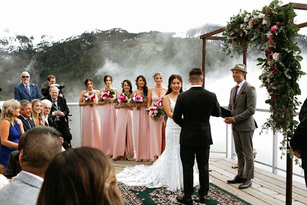 A bride and groom exchange vows outdoors on a mountain deck in Whistler, surrounded by bridesmaids in pink dresses, guests, and a floral arch, with misty mountains beyond—a perfect scene for those planning a wedding in Whistler.