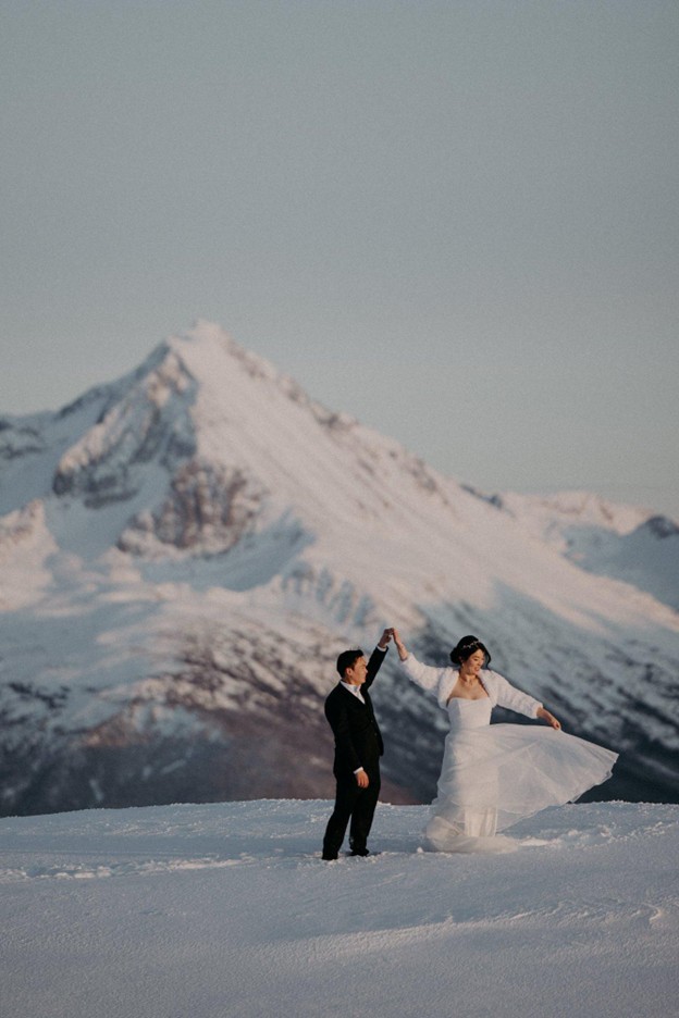 A couple in wedding attire dances on a snowy landscape with a large, snow-covered mountain in the background, capturing the magic of planning a wedding in Whistler.