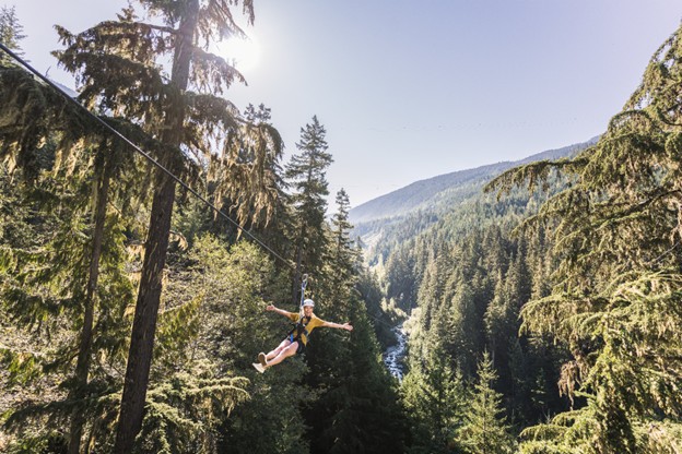A person rides a zipline through a forested mountain landscape on a sunny day, enjoying an alpine adventure with tall trees and a scenic valley—one of the top things to do during Whistler summer.