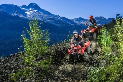 Two people ride red ATVs down a rocky mountain trail with trees and distant snow-capped peaks in the background under a clear sky, capturing the thrill of summer in Whistler.