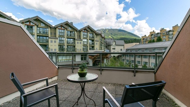 A small outdoor balcony with two chairs and a table overlooks modern buildings and a mountain, perfect for enjoying summer in Whistler under a partly cloudy sky.
