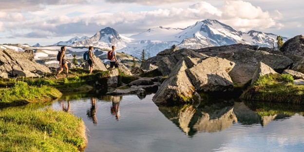 Three hikers walk beside a small pond in a rocky alpine landscape, snow-capped mountains in the background, capturing the essence of summer in Whistler.
