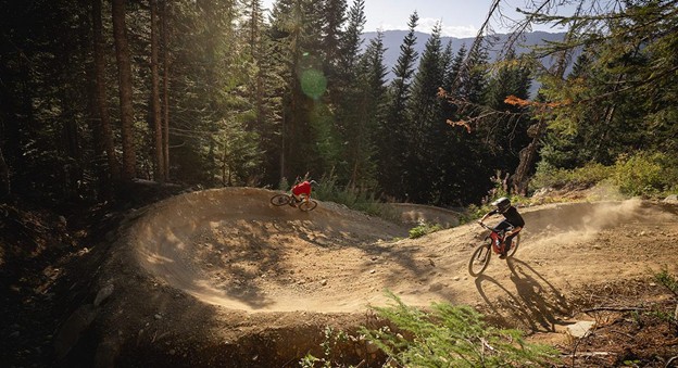 Two mountain bikers ride down a dirt trail with banked turns through a forested area, soaking up the sun during summer in Whistler.