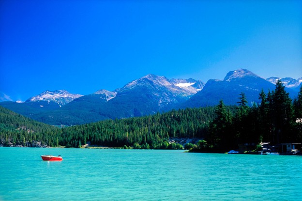 A turquoise lake with a red boat in the water, surrounded by pine trees and mountains under a clear blue sky—an idyllic scene of summer in Whistler.