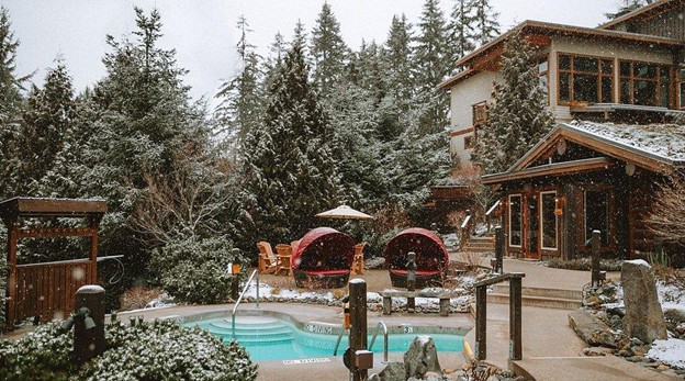 Outdoor hot tub and lounge area at a lodge-style building, surrounded by snow-covered trees and light snowfall—a serene contrast to the lively summer in Whistler.