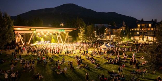 Outdoor concert at dusk during summer in Whistler, with a crowd gathered near a lit stage, people relaxing on the grass, and majestic mountains in the background.