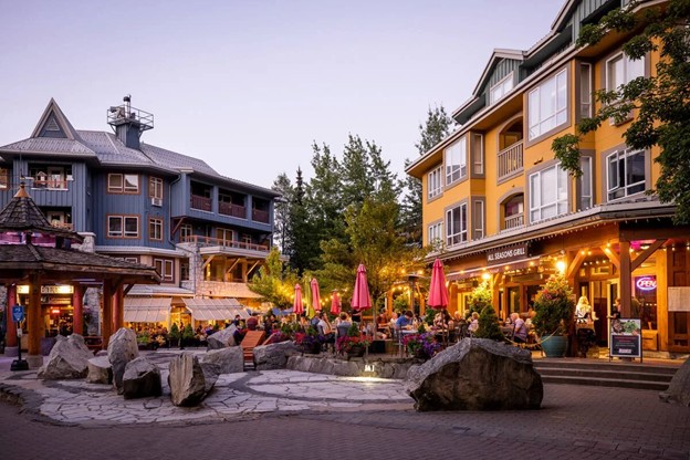 Outdoor dining area with pink umbrellas and people seated at tables beside colorful buildings and large rocks in a pedestrian plaza at dusk, capturing the vibrant atmosphere of summer in Whistler.